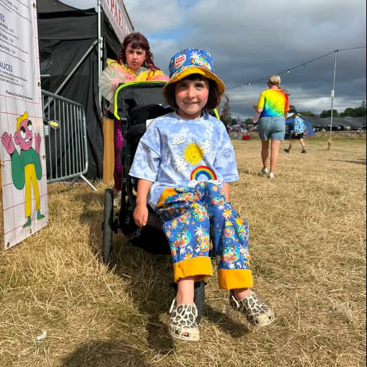 A child wearing a blue sun graphic t-shirt, colorful patterned pants, and a matching bucket hat sits in a stroller at an outdoor festival on a grassy field.