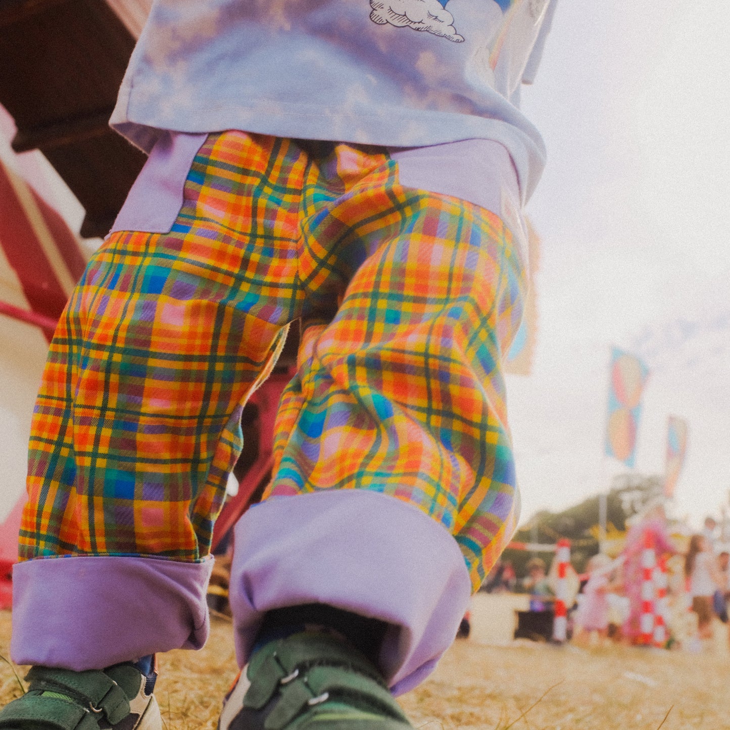 A close-up lifestyle shot of a child's legs wearing colorful plaid pants with purple cuffs, standing outdoors at what looks like an outdoor festival or fair.