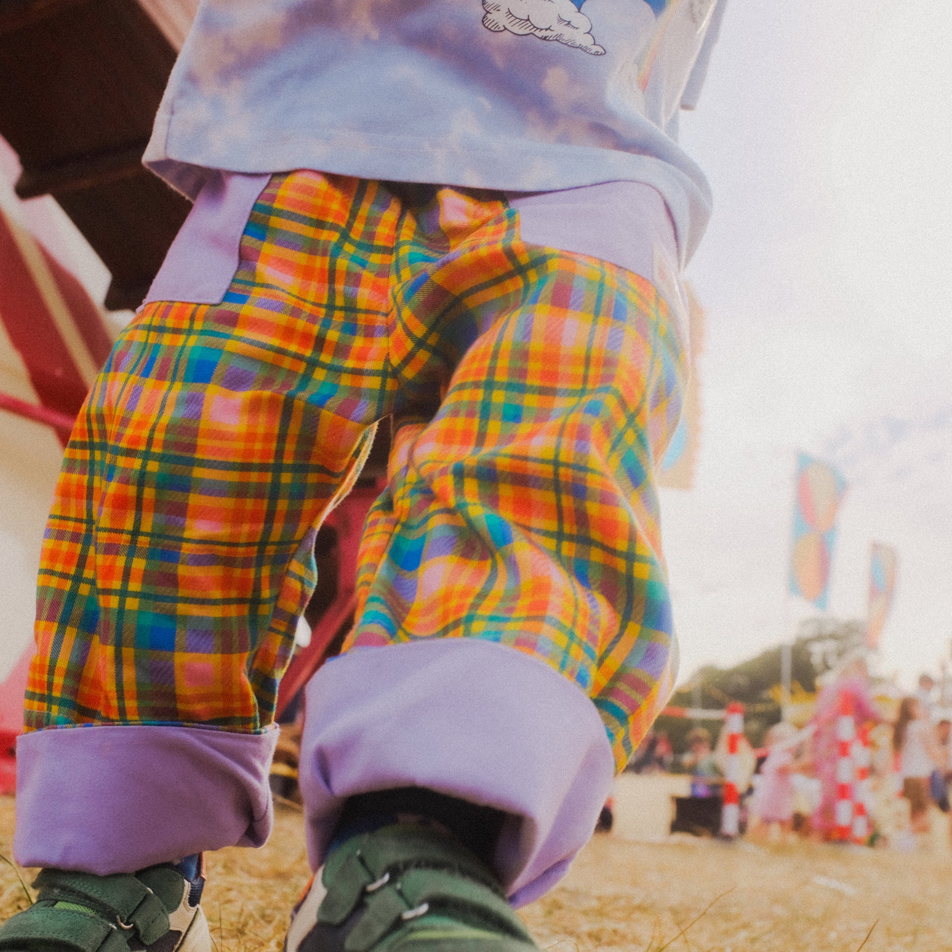 A close-up lifestyle shot of a child's legs wearing colorful plaid pants with purple cuffs, standing outdoors at what looks like an outdoor festival or fair.