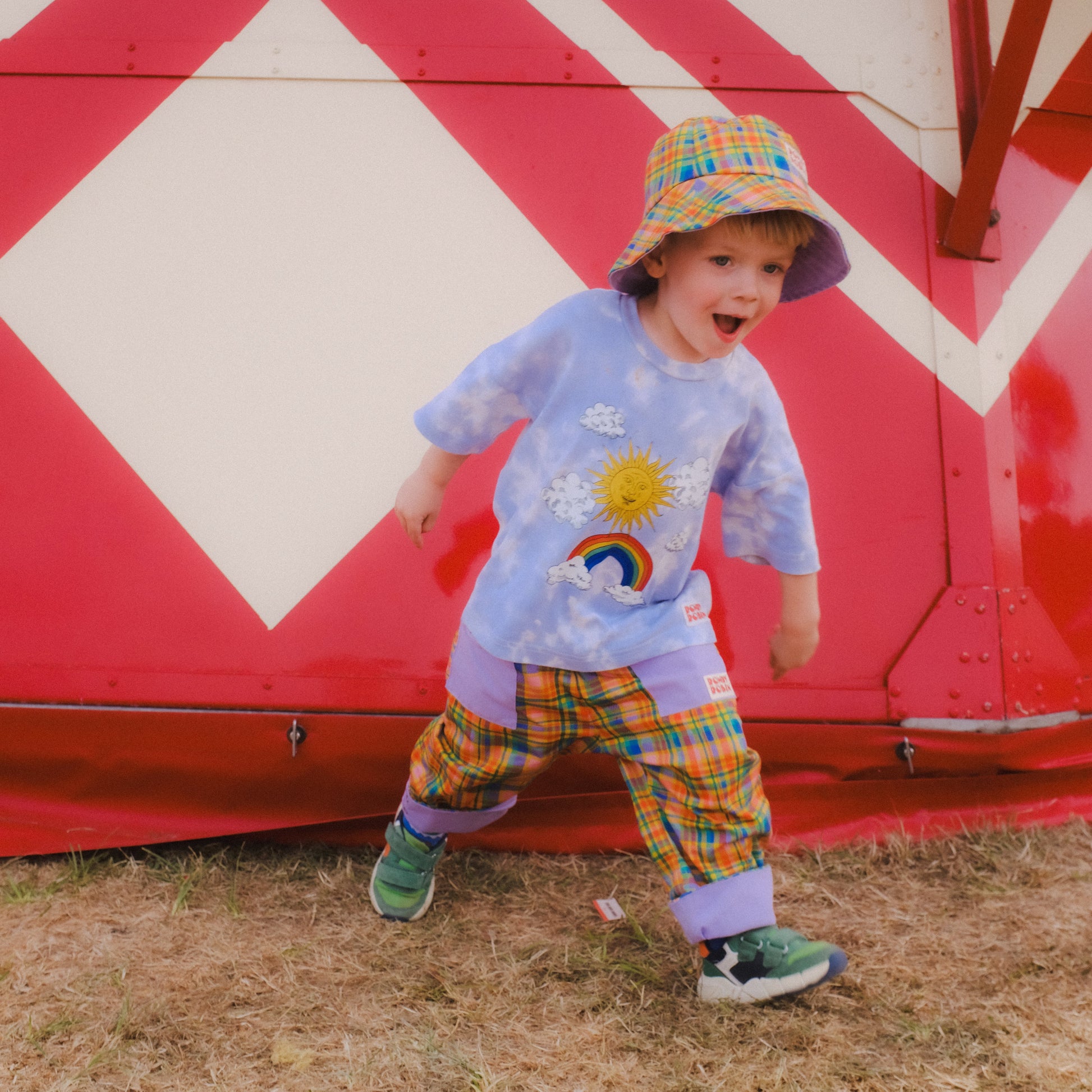 A toddler wearing a colorful plaid bucket hat, a light blue tie-dye t-shirt with a sun and rainbow graphic, and matching plaid pants stands in front of a red and white striped trailer.