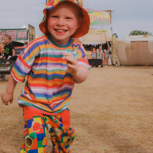 A young child wearing a colorful rainbow-striped t-shirt, matching patterned pants, and a bucket hat, smiling outdoors on dry grass with a fairground or market in the background.