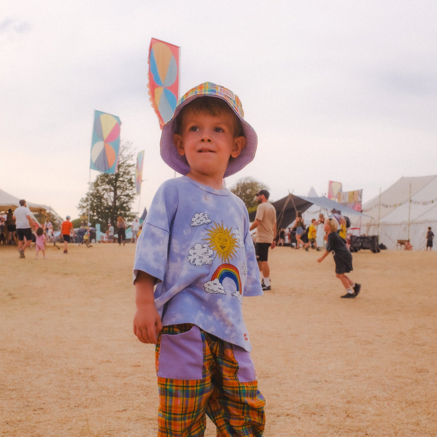 A young child standing outdoors on a sandy field at what appears to be a fair or festival, wearing a lavender bucket hat, a light purple graphic T-shirt, purple plaid pants, and green sneakers.