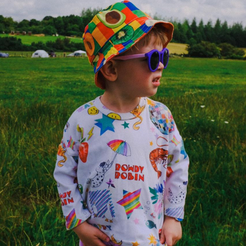 A young child wearing a colorful patchwork bucket hat, purple sunglasses, a white long-sleeve shirt with a vibrant rainbow space-themed print, and lavender pants, standing in a grassy field under a cloudy sky.